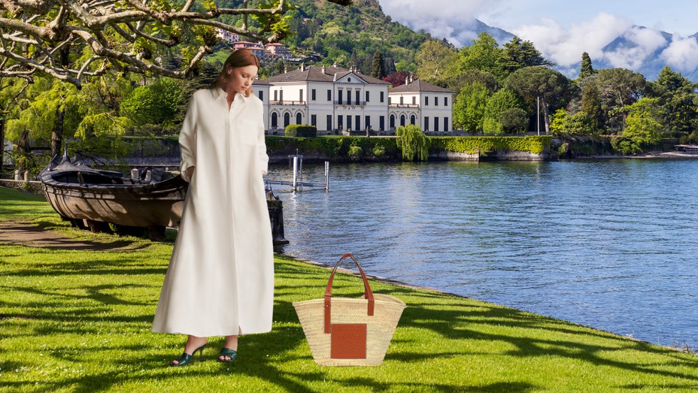 woman in stylish white dress standing on grass by Lake Como with a straw bag