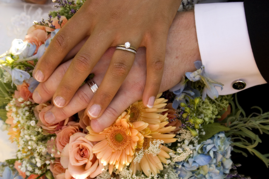 Two different hands are clasped together, resting on a bouquet of flowers, showing their wedding rings.
