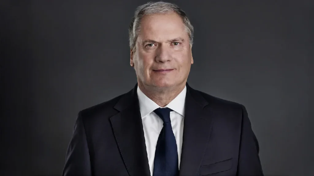 A white older male in a black suit with a dark background.