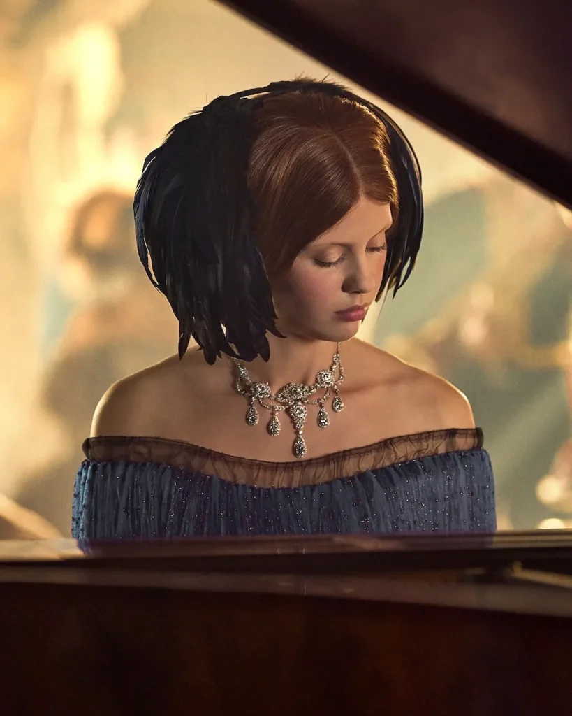 A white woman in period attire sits at a piano with a dazzling diamond necklace around her neck.
